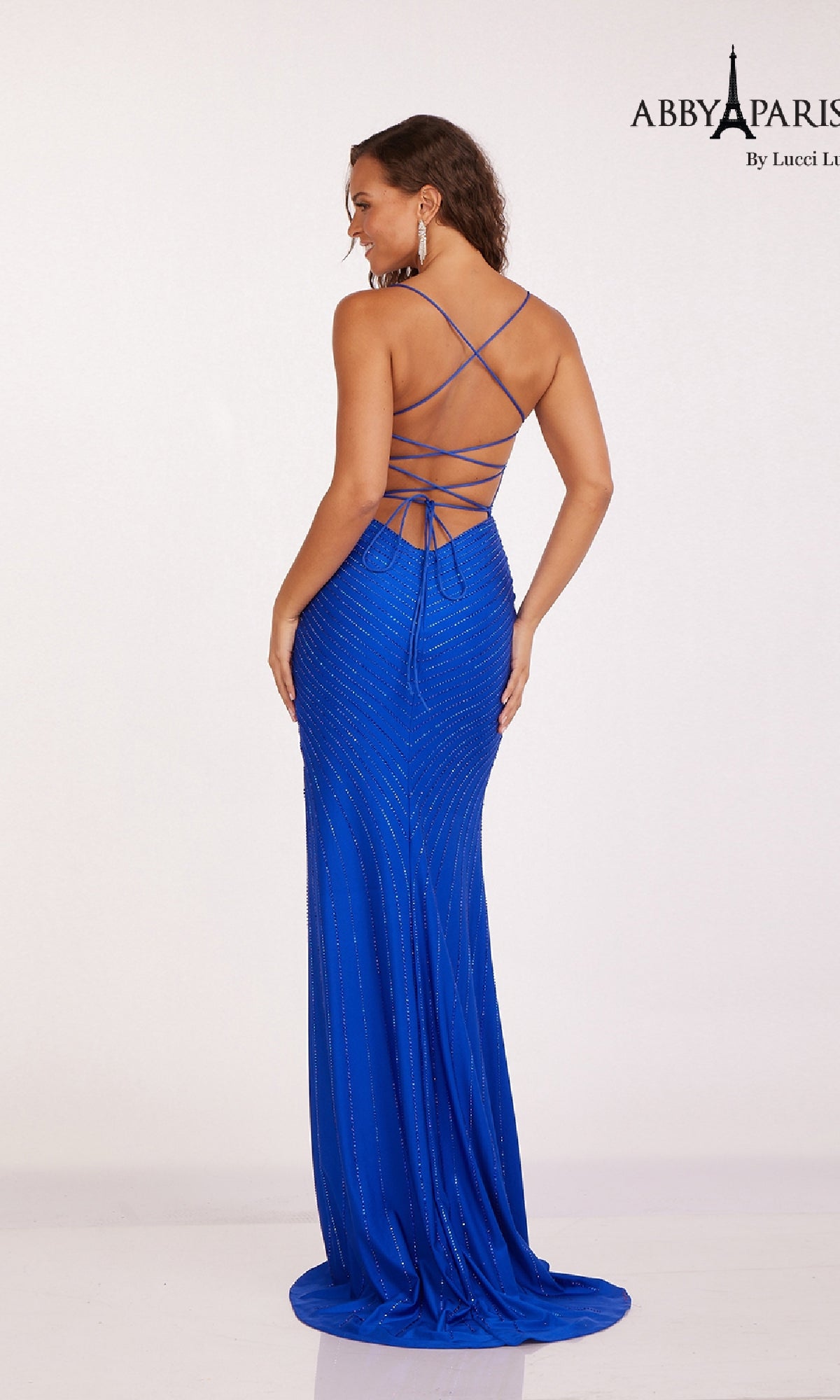 A woman models the Abby Paris 90201 Empire-Waist Long Prom Dress—bright blue with thin straps and an open crisscross back—against a plain light background.